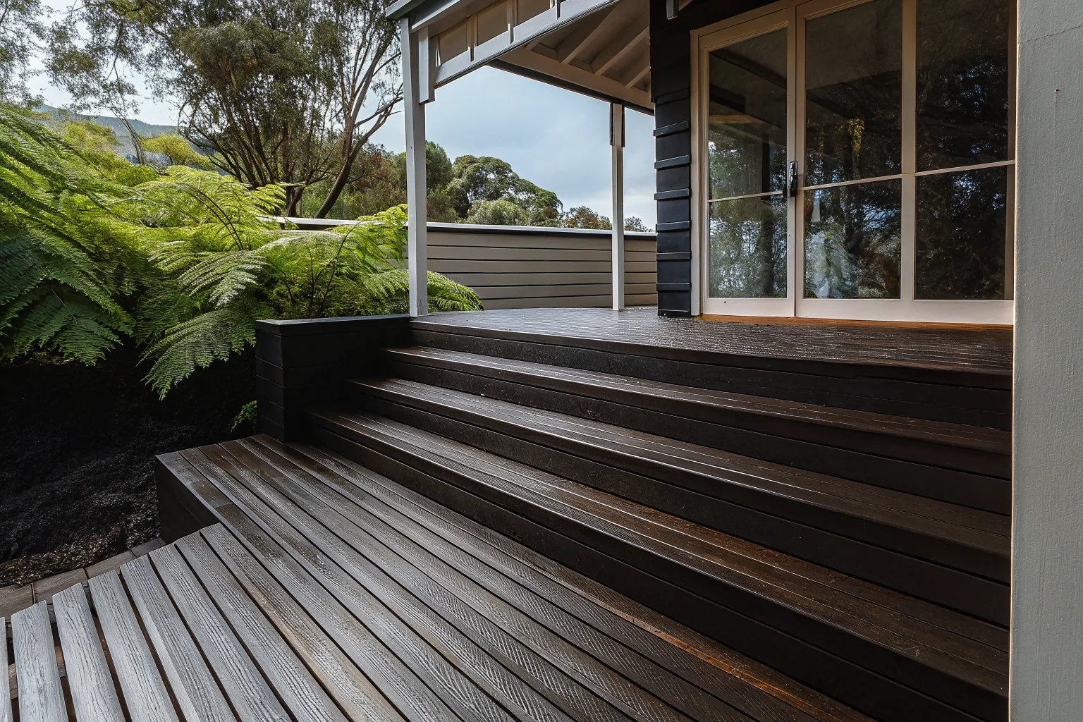 Dark-stained timber deck with steps leading up to a home surrounded by ferns